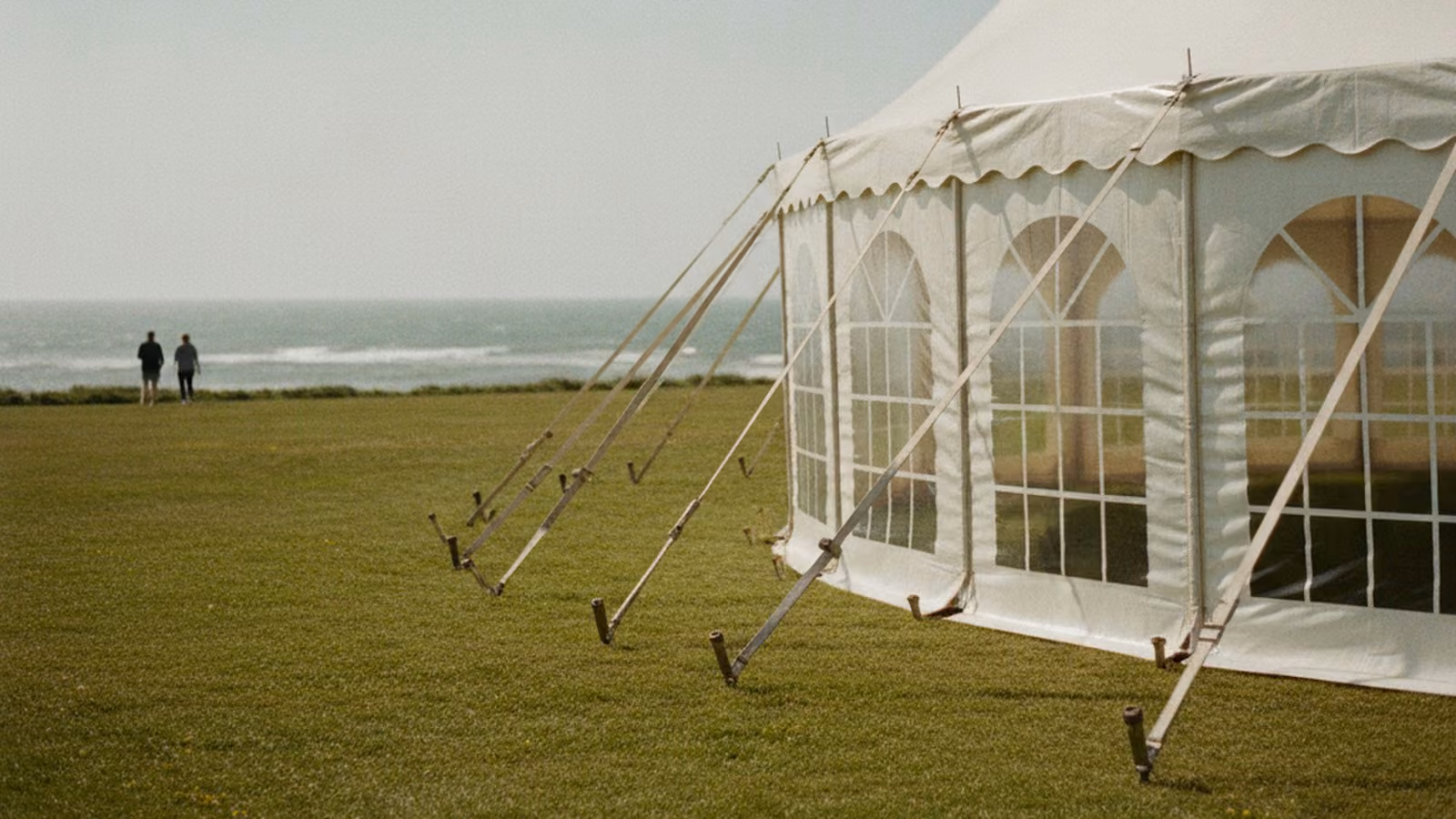 A white event tent on a coastal lawn with guy ropes taut in the wind